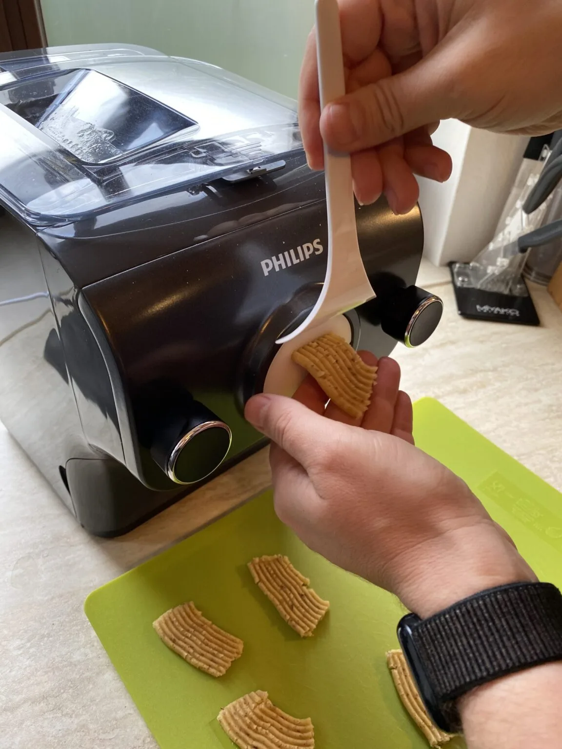 Aus dem Philips Pastamaker kommt gerippter Teig, den eine Hand abschneidet; fertige Teigstücke liegen auf grünem Schneidebrett.