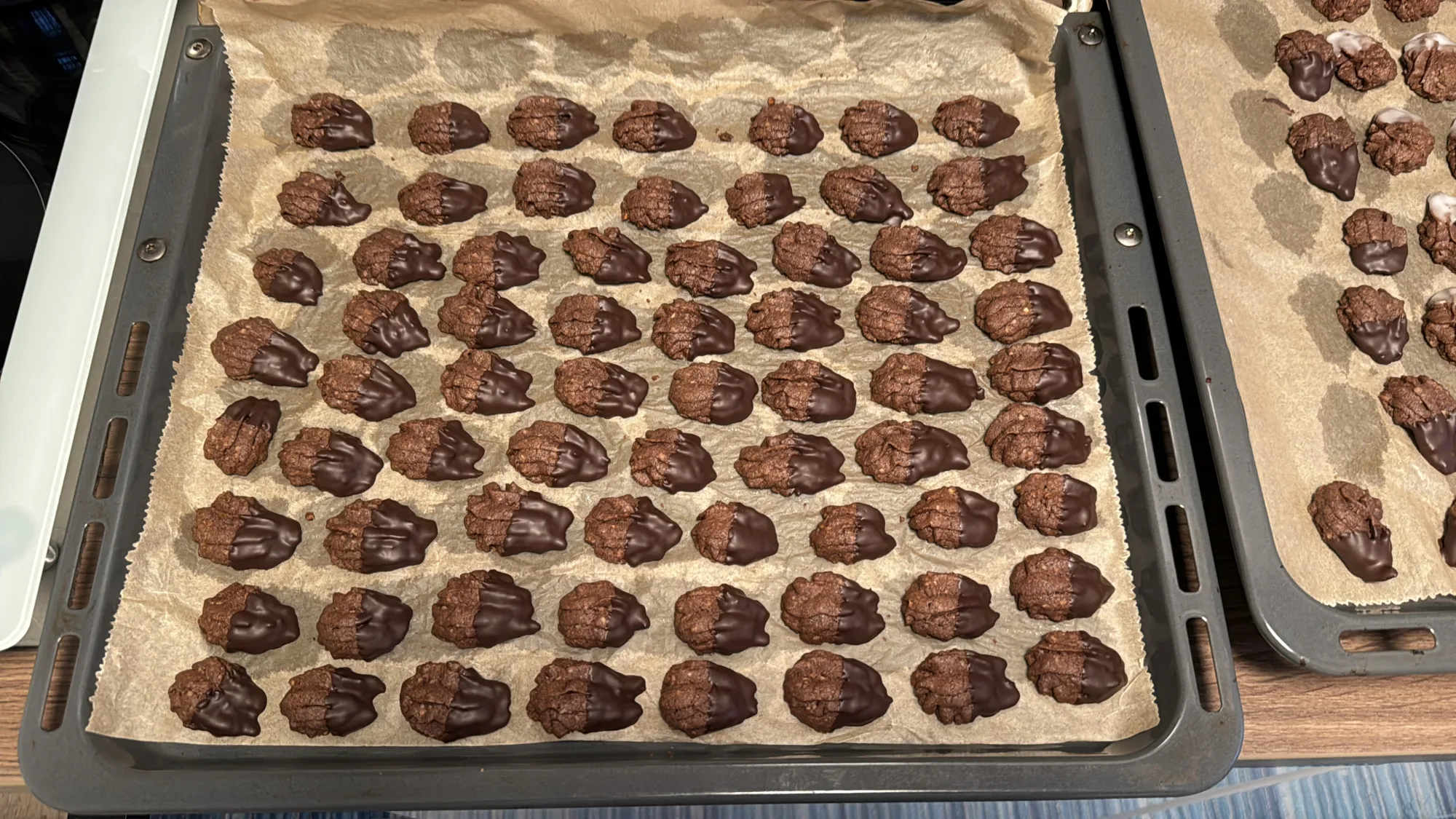 Backblech mit Backpapier und vielen kleinen Schokoladenplätzchen, deren eine Hälfte in dunkle Kuvertüre getaucht ist