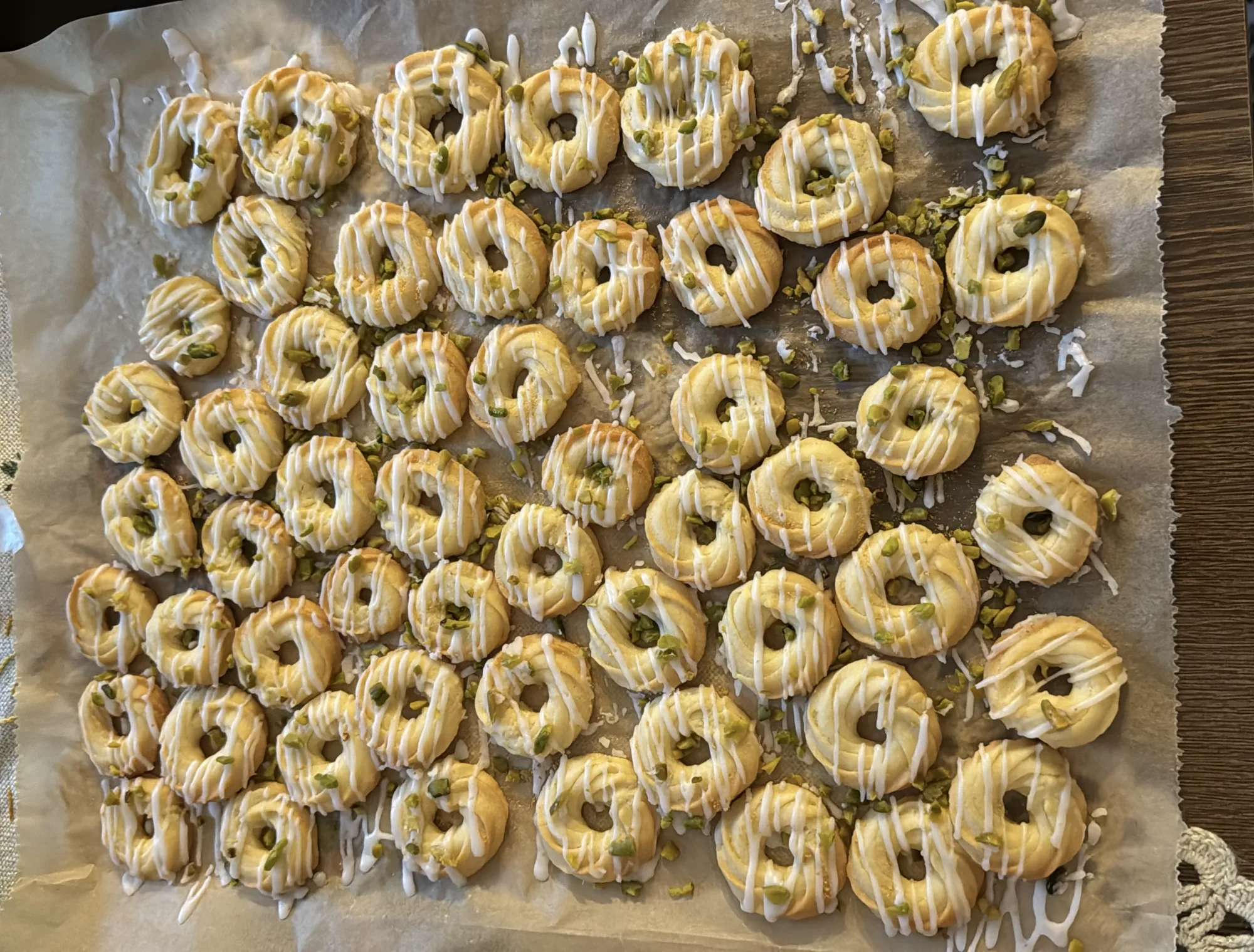 Spritzgebäck-Ringe auf Backpapier, mit weißen Zuckerglasur-Streifen und gehackten Pistazien bestreut, auf einem Holztisch.