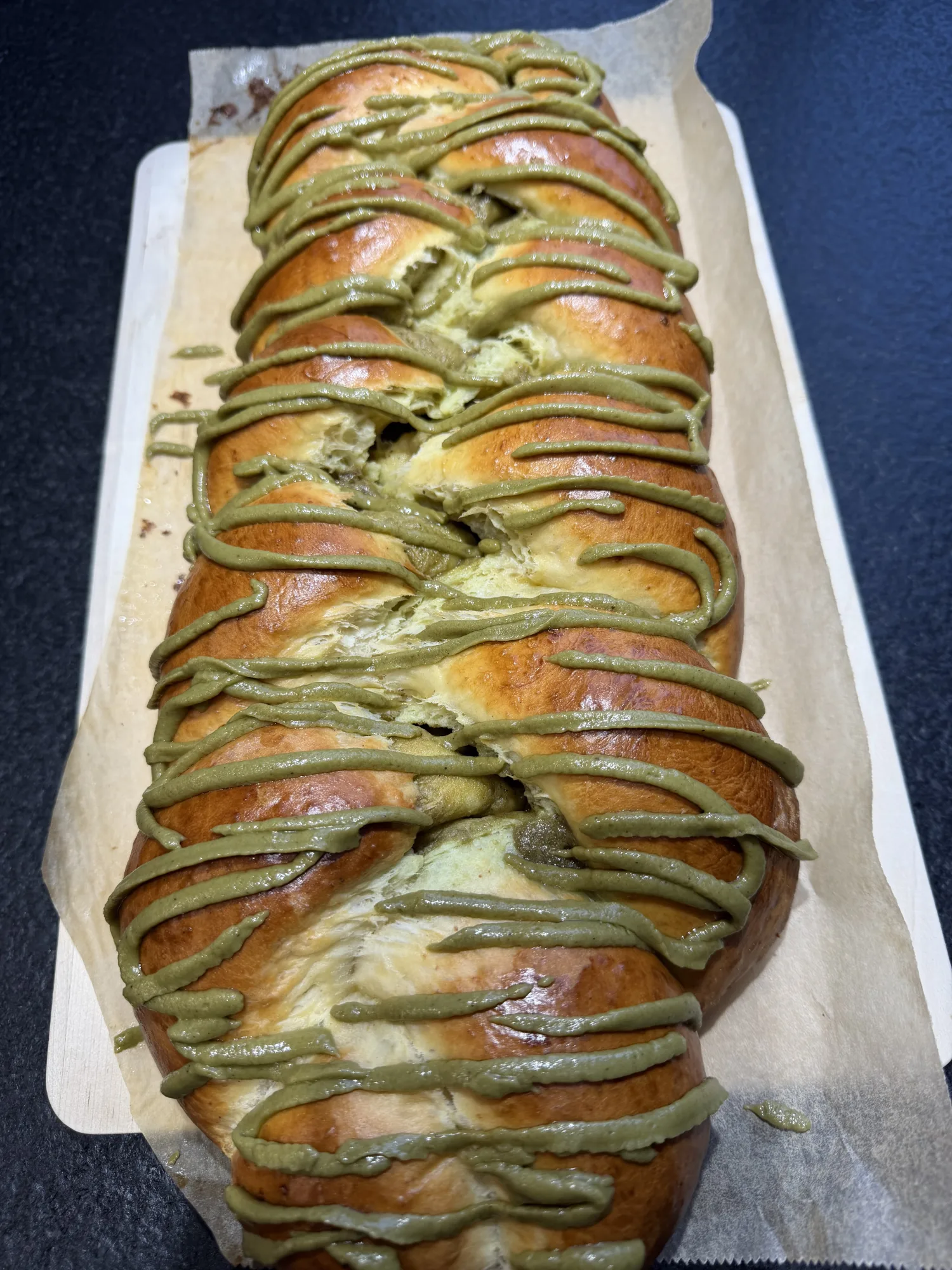 Goldbrauner Hefezopf mit grünen Pistaziencreme-Streifen, auf Backpapier und einem Holzbrett, aufgerissene Stellen zeigen den Teig.