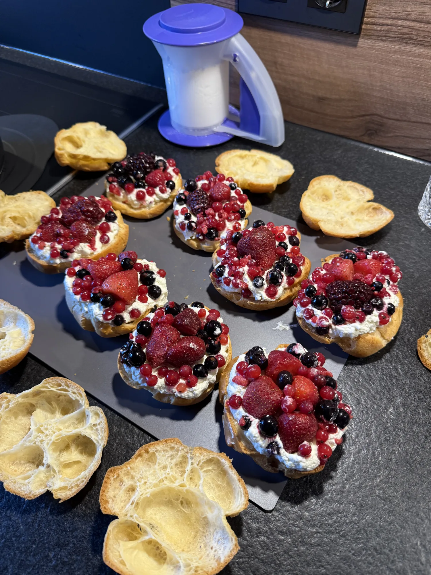 Windbeutel mit Sahne und gemischten Beeren auf einem Blech, daneben abgenommene hohle Brandteig-Deckel und ein Puderzuckerstreuer.