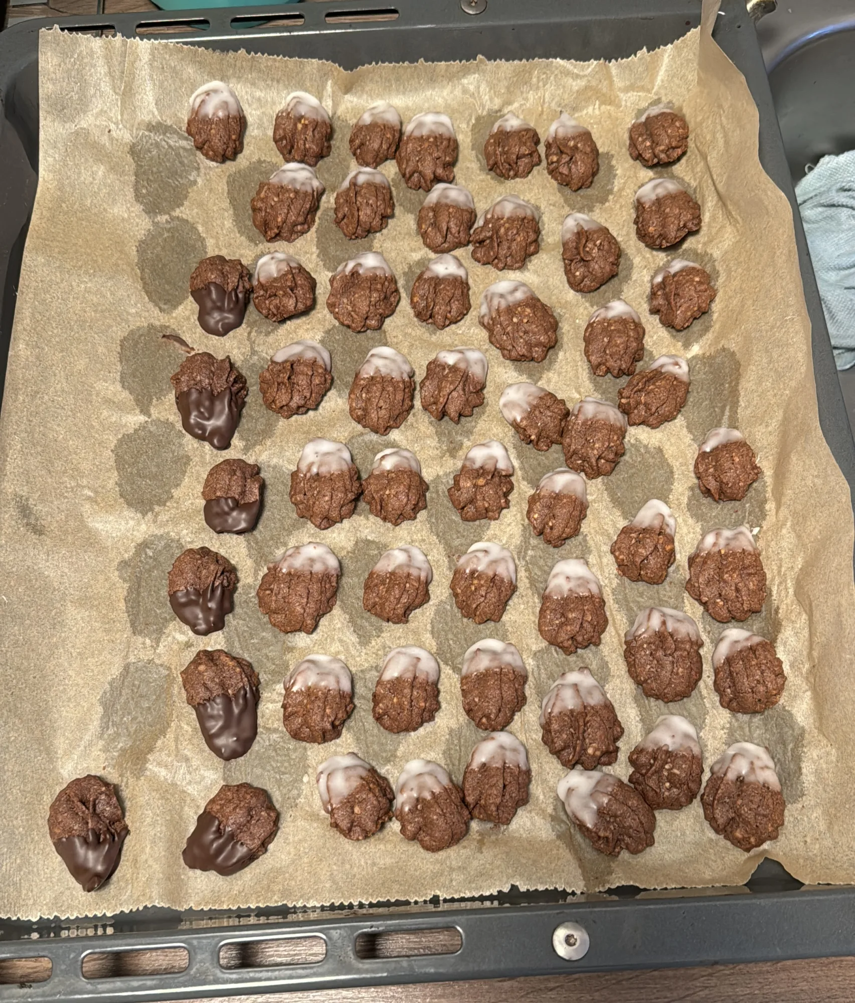Kleine dunkle Gebäckhäufchen auf Backpapier am Blech, die meisten mit weißer Zuckerglasur, einige mit Schokolade an einem Ende