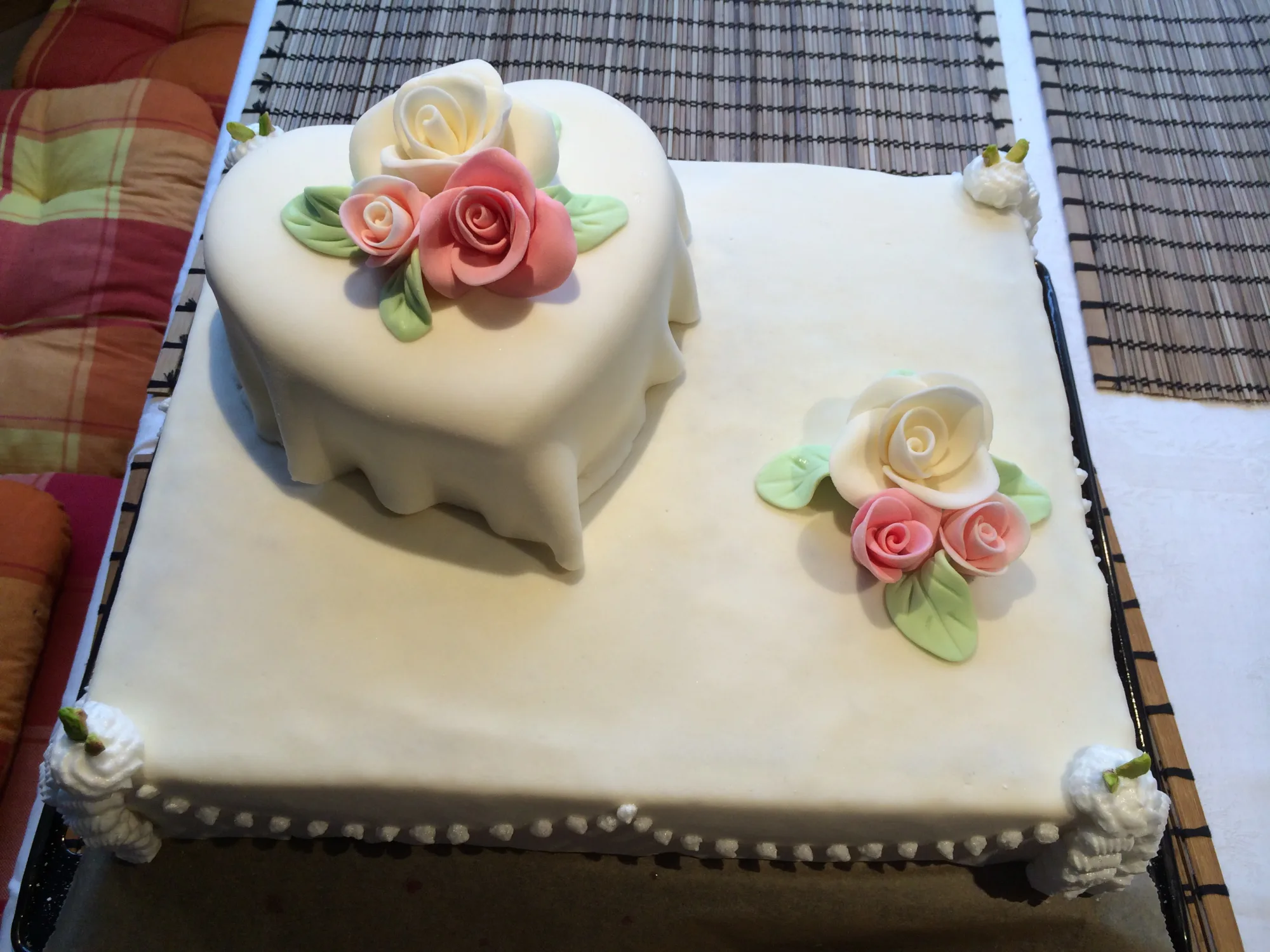Hochzeitstorte mit weißem Fondant: herzförmige Torte auf eckigem Blechkuchen, dazu rosa und weiße Zuckerrosen und Spritzbeutel-Ecken.