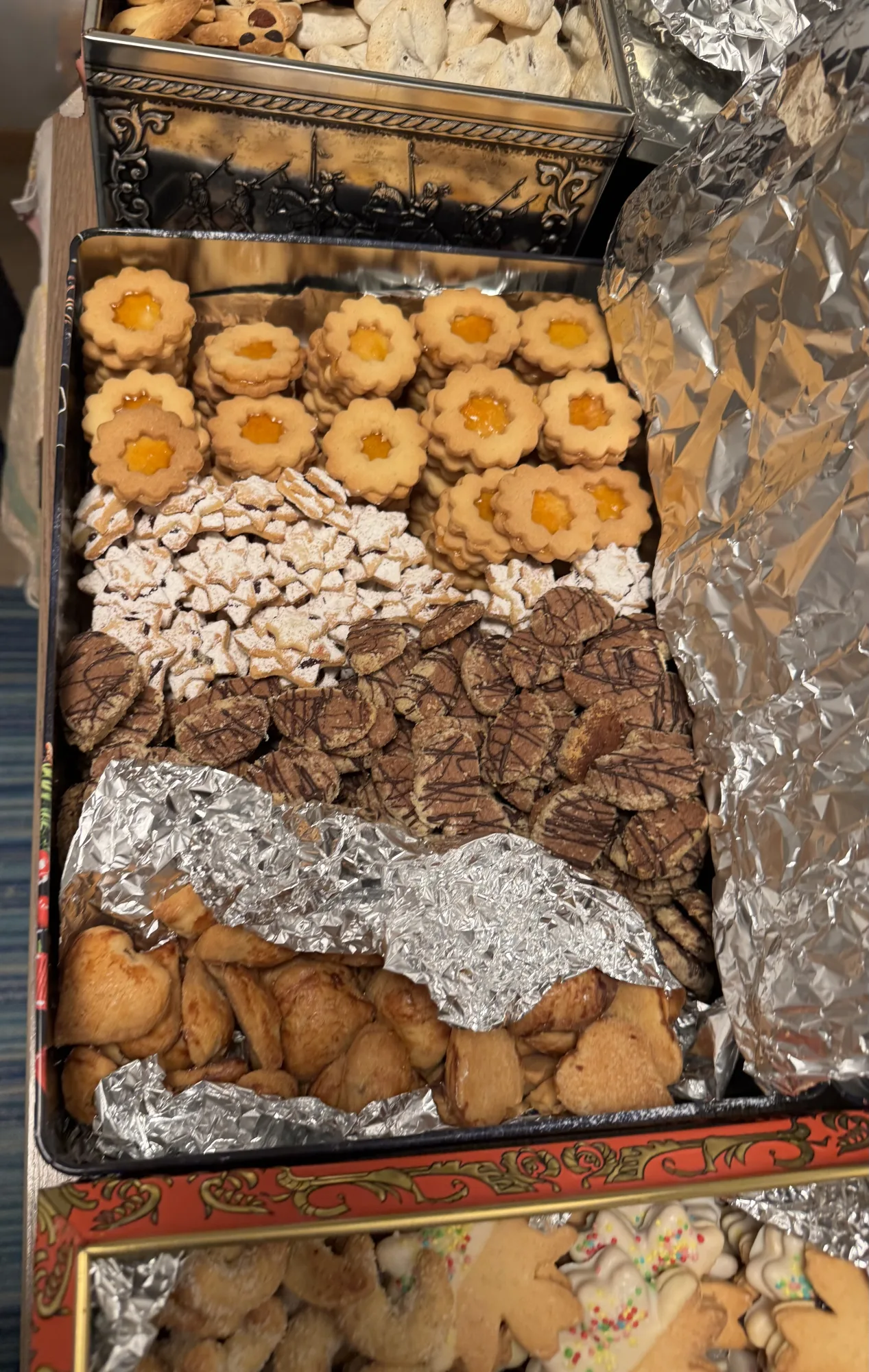 Plätzchendose mit Linzer Augen, Puderzucker-Sternen, schokoladenbestreiften Keksen und Hörnchen, getrennt durch Alufolie.