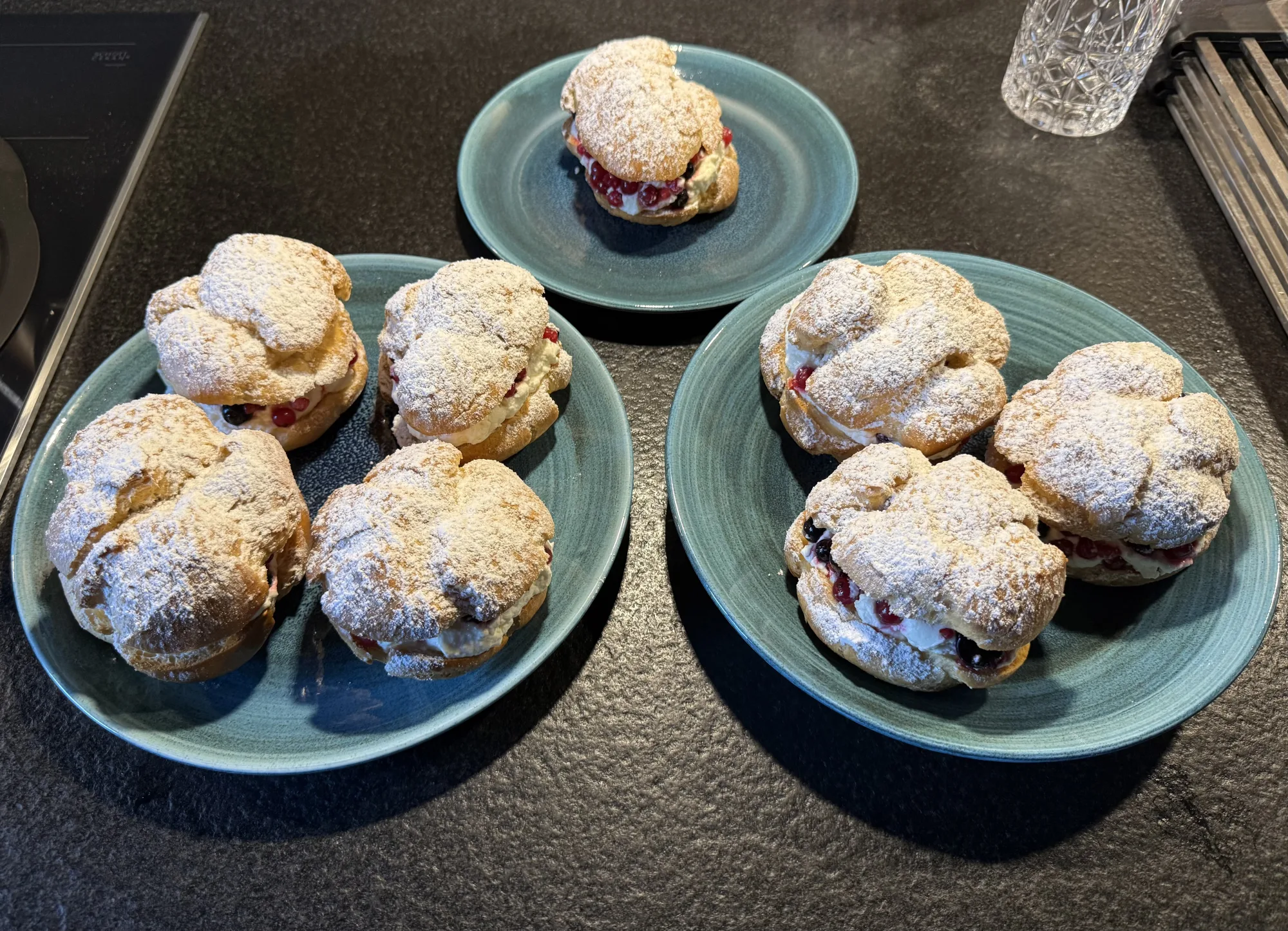 Sieben Windbeutel mit Puderzucker, Sahne und roten sowie schwarzen Johannisbeeren auf drei blaugrünen Tellern, Arbeitsplatte dunkel.
