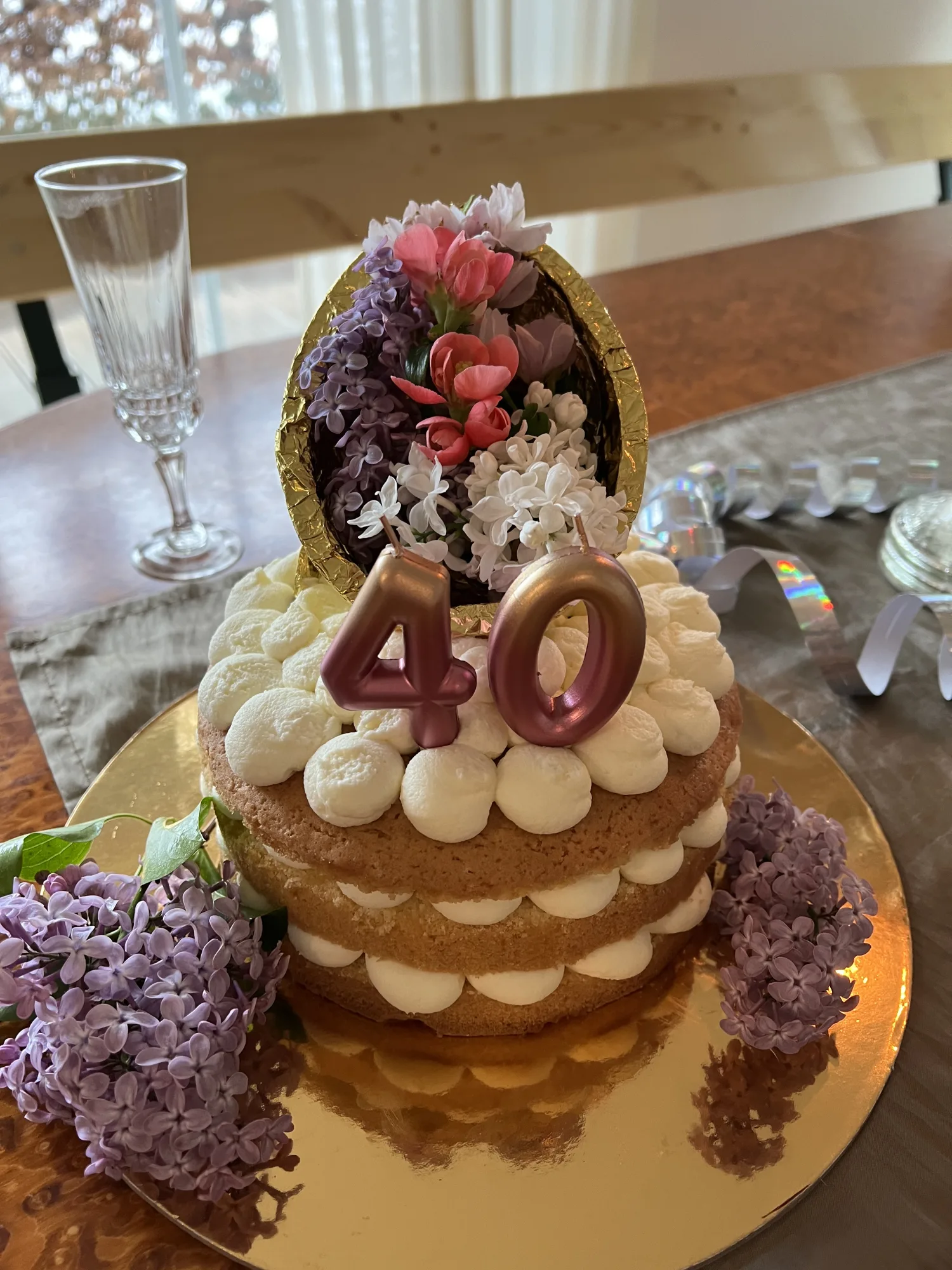 Dreistöckige Naked Cake mit Sahnetupfen, Kerzen "40" und goldener Schale voller Flieder- und Zierquittenblüten auf Goldplatte