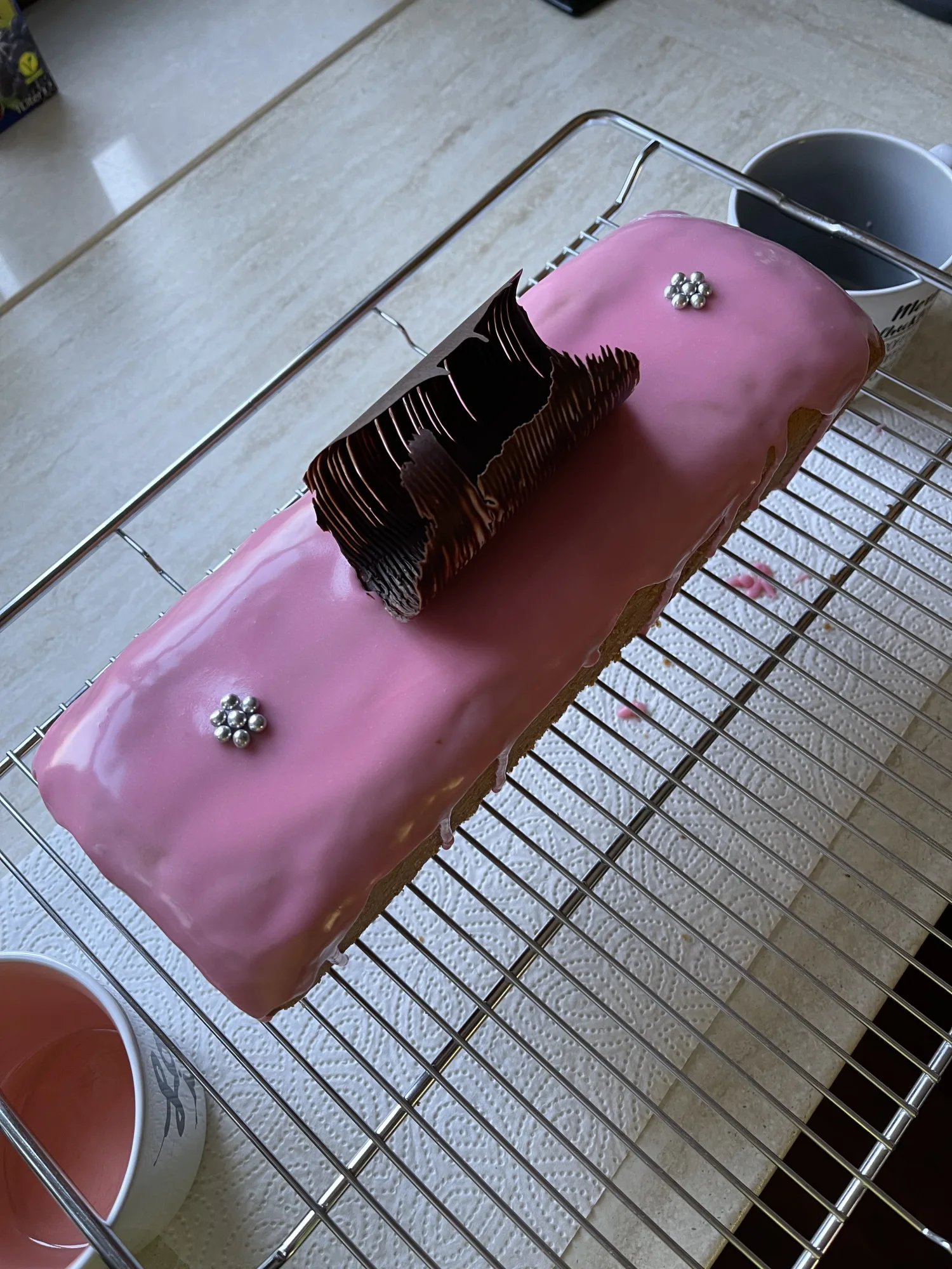 Kastenkuchen mit rosa Glasur, Schokoladenzier und zwei Silberperlen-Blümchen, auf einem Kuchengitter über Küchenpapier.
