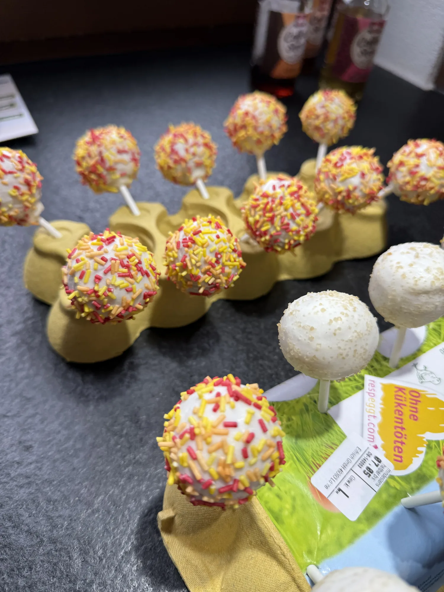 Cakepops mit weißer Glasur, bunten Zuckerstreuseln und Zuckerkristallen, in Eierkartons auf einer dunklen Schieferplatte gesteckt