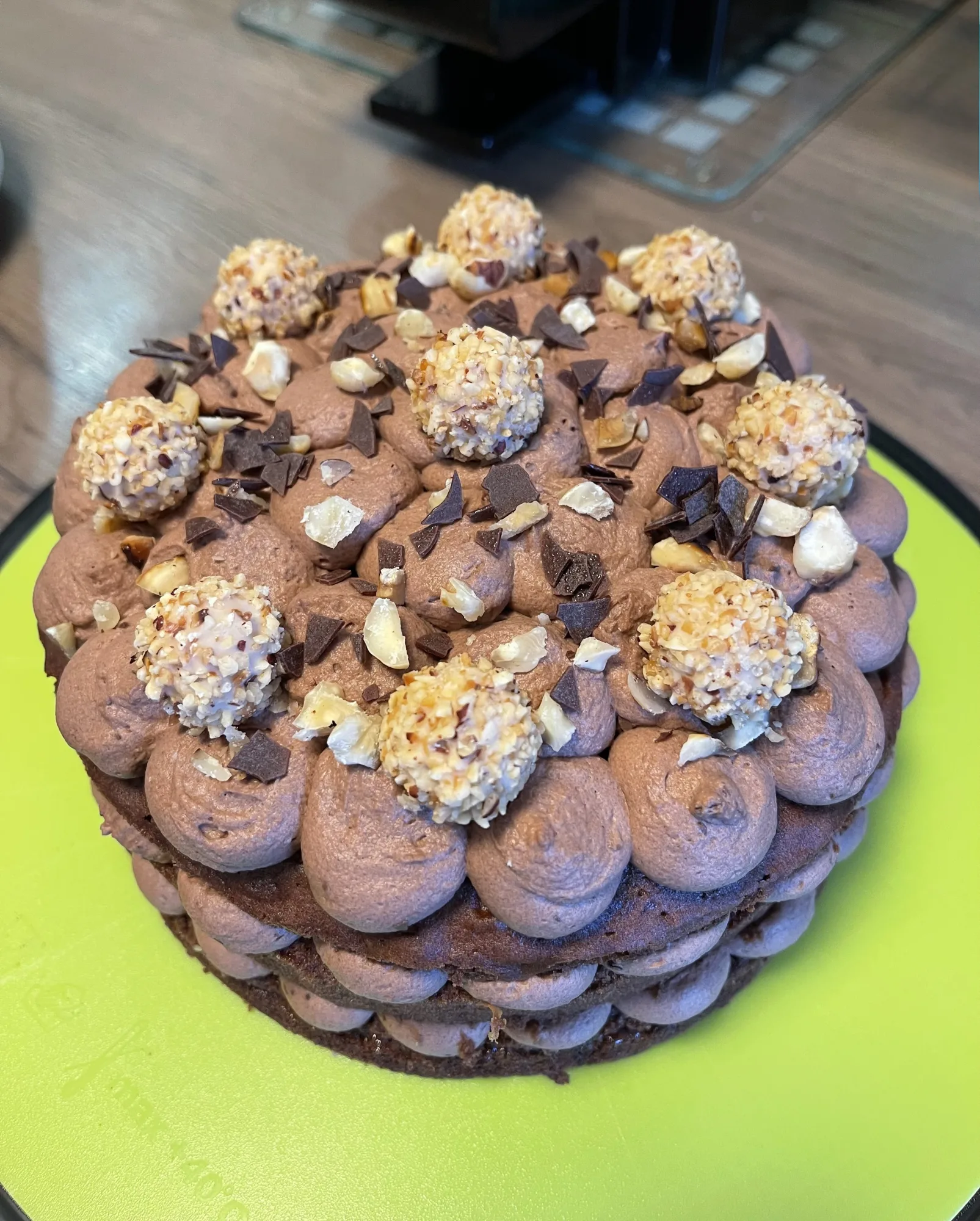 Kleine Torte mit Schokoladenböden, getupfter Nugatcreme, Haselnusskrokant-Kugeln, Schokospänen und Nussstücken auf grüner Platte