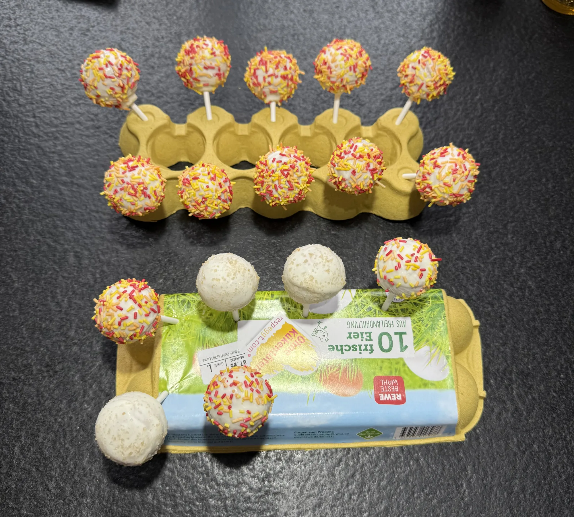 Cakepops mit weißer Glasur, die meisten mit rot-gelb-orangen Streuseln, stecken in Eierkartons auf dunkler Arbeitsfläche.