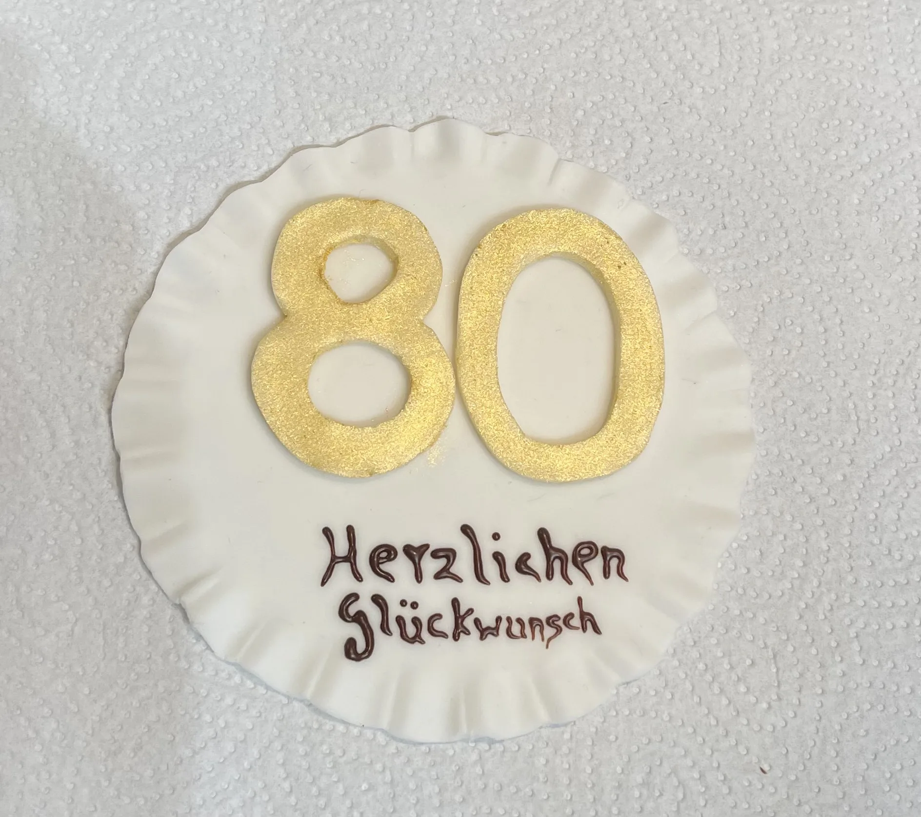 Weiße Fondantscheibe mit Rüschenrand, goldener Zahl 80 und braunem Schokoschriftzug „Herzlichen Glückwunsch“ auf Papierserviette