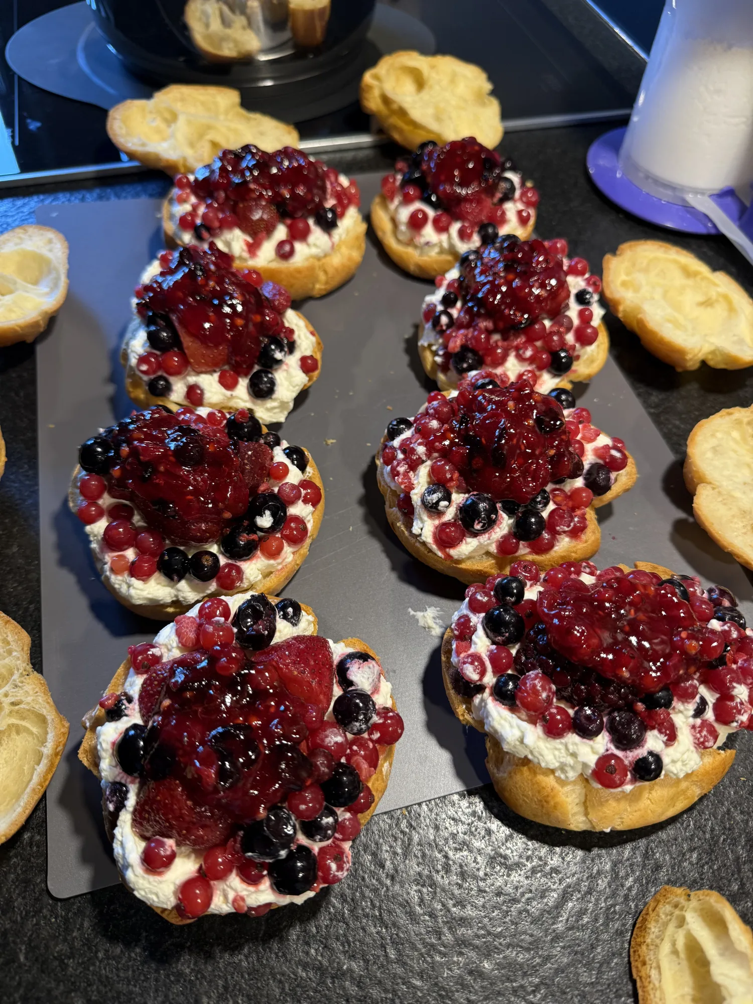 Windbeutel-Böden mit Sahnecreme, roten und schwarzen Johannisbeeren und Beerenkompott auf einem Backblech, daneben die Deckel