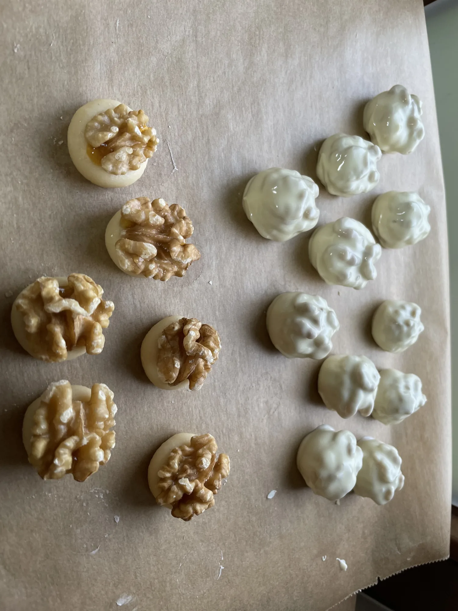 Kleine runde Mürbteigplätzchen mit Walnusshälften links, rechts weiß glasierte Walnuss-Plätzchen, auf Backpapier.