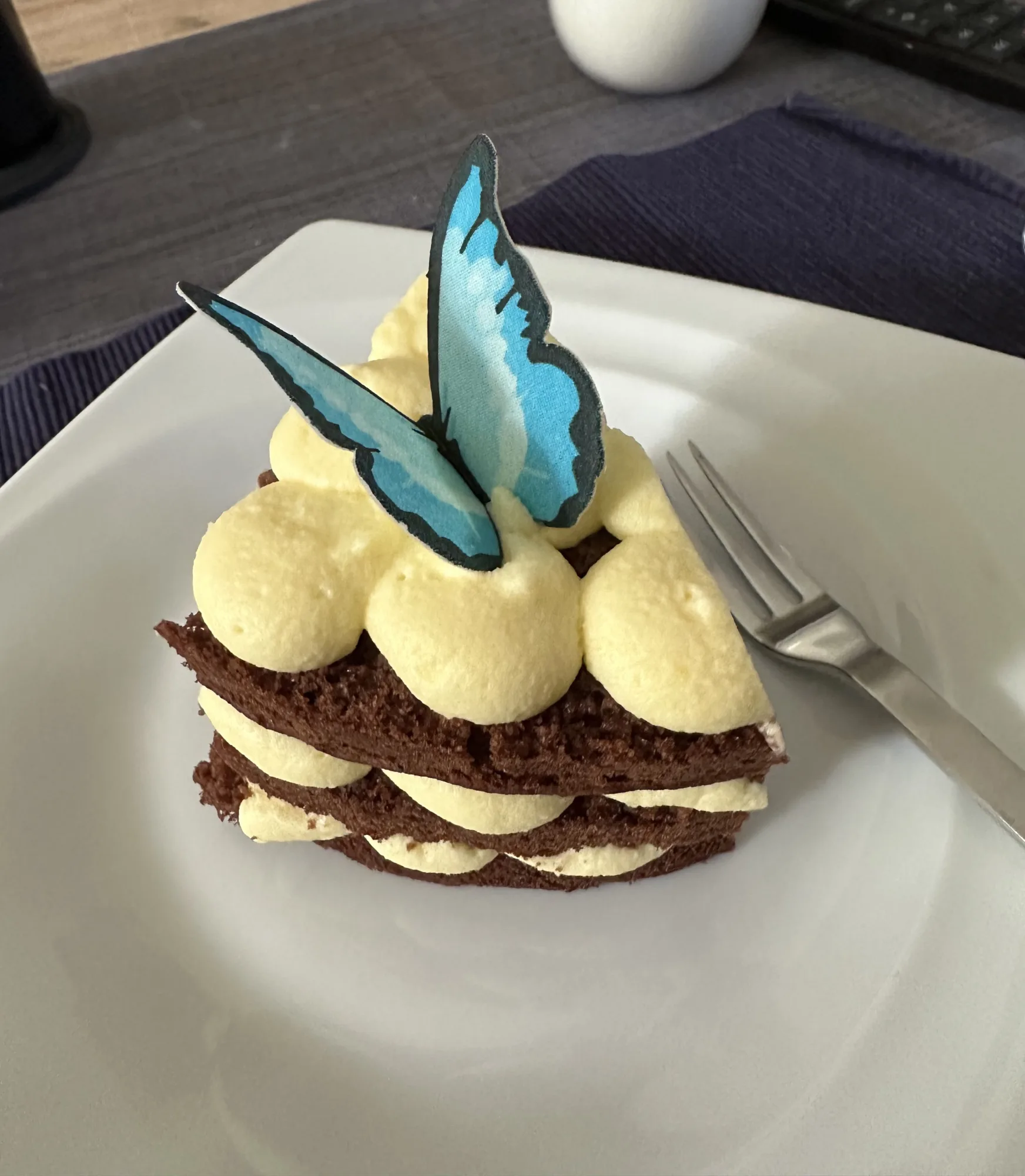Kleine Torte mit Schokoladenbiskuit, gelben Cremetupfen und einem blauen Schmetterling als Deko auf weißem Teller mit Gabel