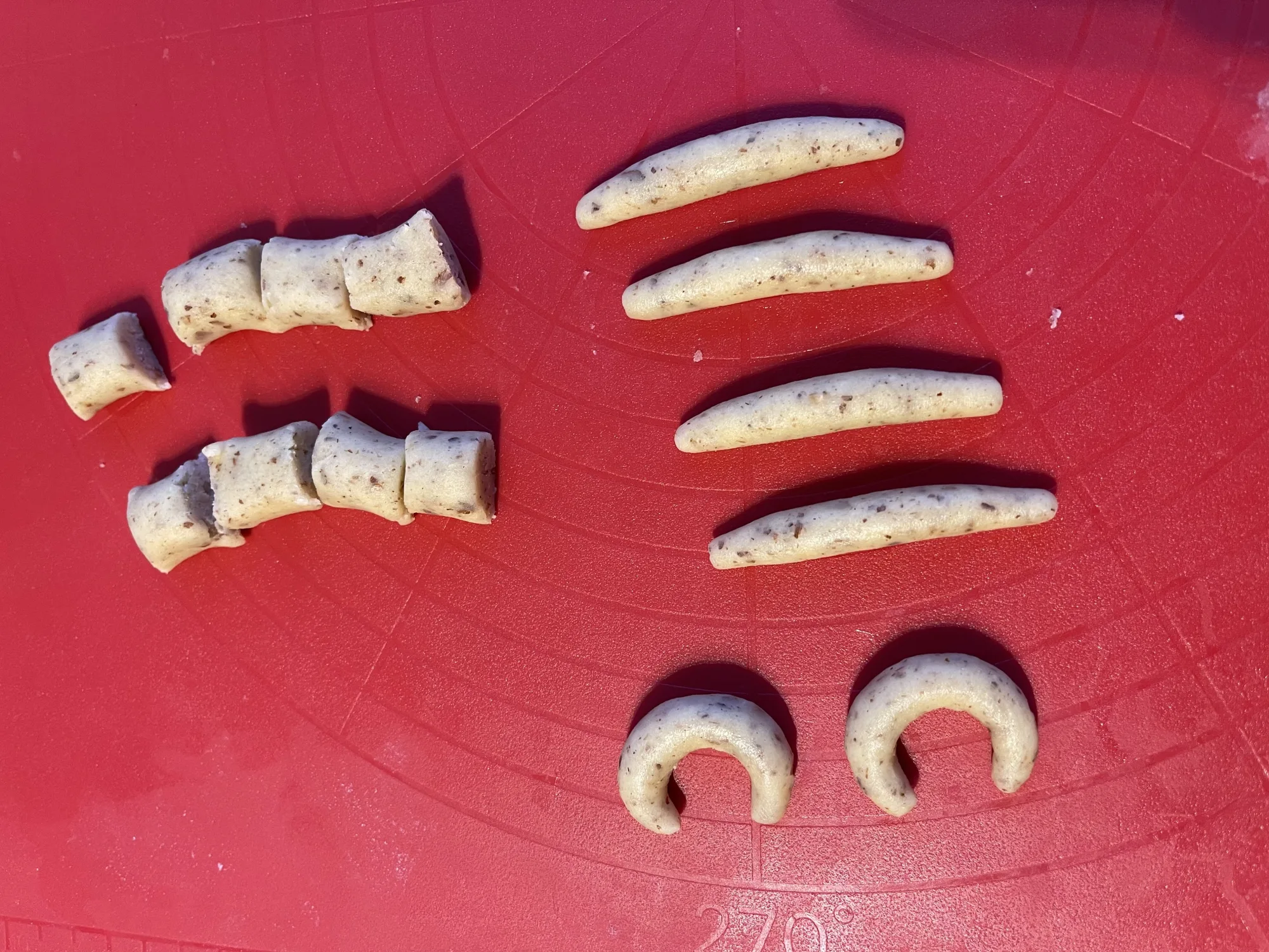 Teigstücke, dünne Teigrollen und zwei zu Hörnchen gebogene Teiglinge mit dunklen Sprenkeln auf einer roten Silikon-Backmatte