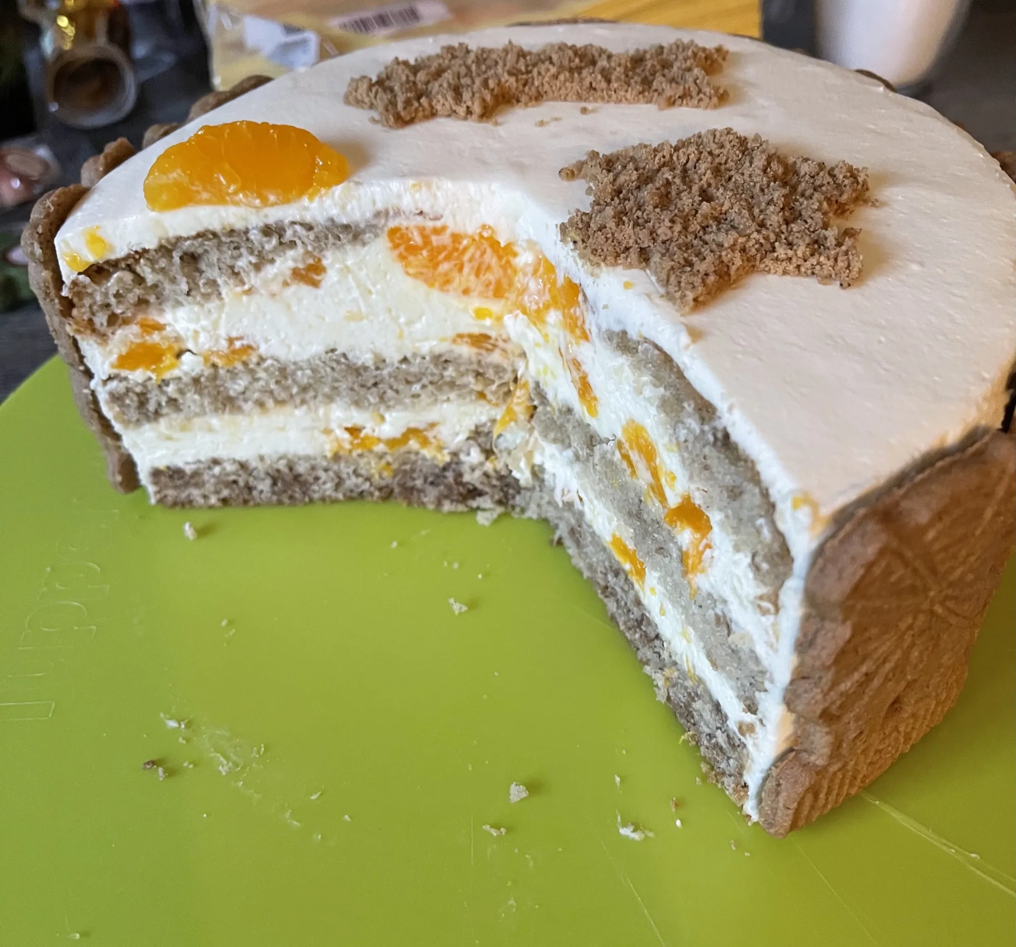 Angeschnittene kleine Torte mit Sahnecreme, Mandarinenstücken, Gewürzbiskuit, Keksrand und Krümeln auf grünem Brett