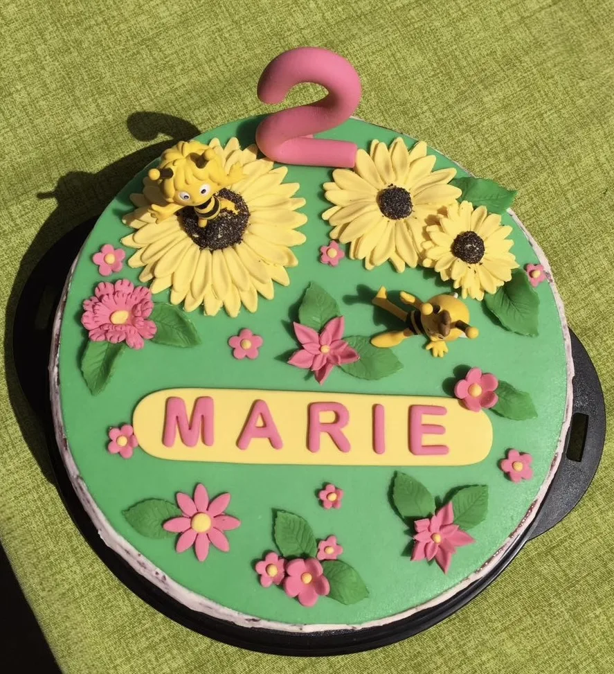 Namenstorte mit grünem Fondant, Sonnenblumen, rosa Blüten, Biene Maja, rosa Zahl 2 und gelbem Namensschild „MARIE“.
