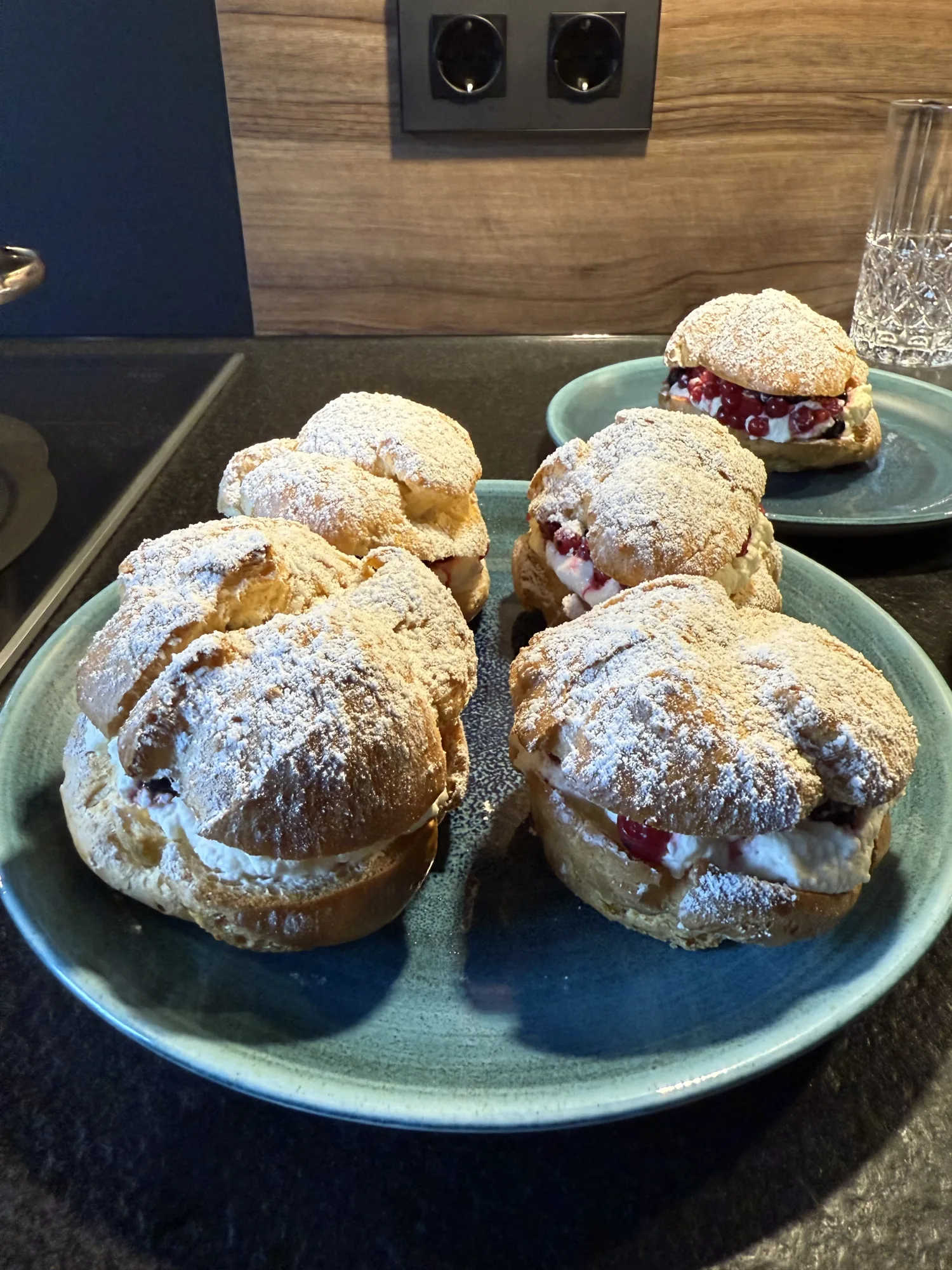 Windbeutel mit Puderzucker, Sahne und roten Johannisbeeren, vier Stück auf einem blaugrünen Teller, einer auf einem zweiten.