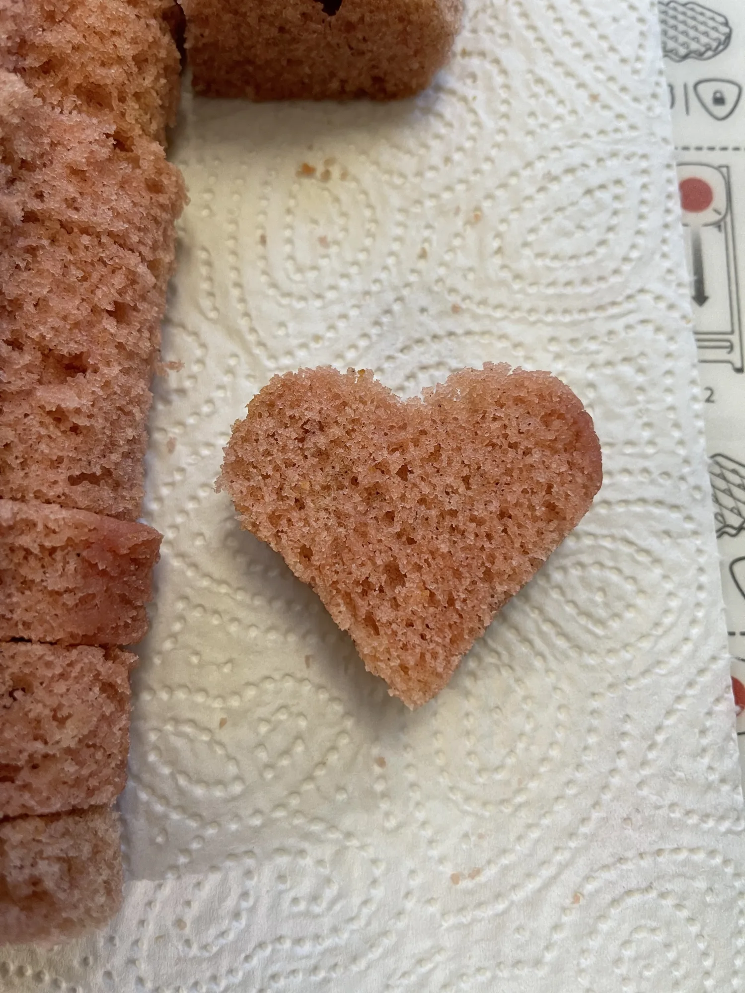 Herzförmig ausgestochenes Stück rosa Rührteig auf Küchenpapier, daneben aufgeschnittene Kuchenscheiben für den Kuchen mit verstecktem Herz