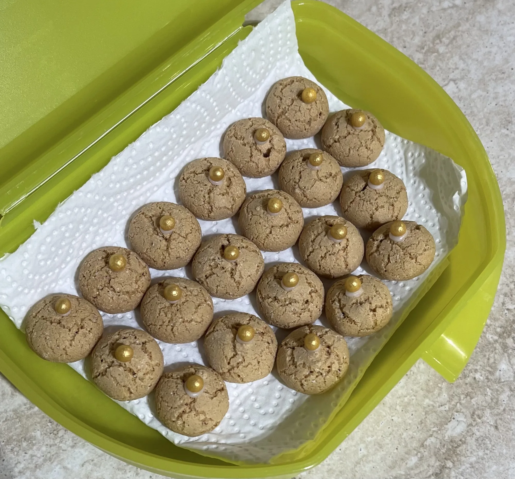 Kleine rissige Nussmakronen mit goldener Zuckerperle obenauf, auf Küchenpapier in einer grünen Plastikdose angeordnet.