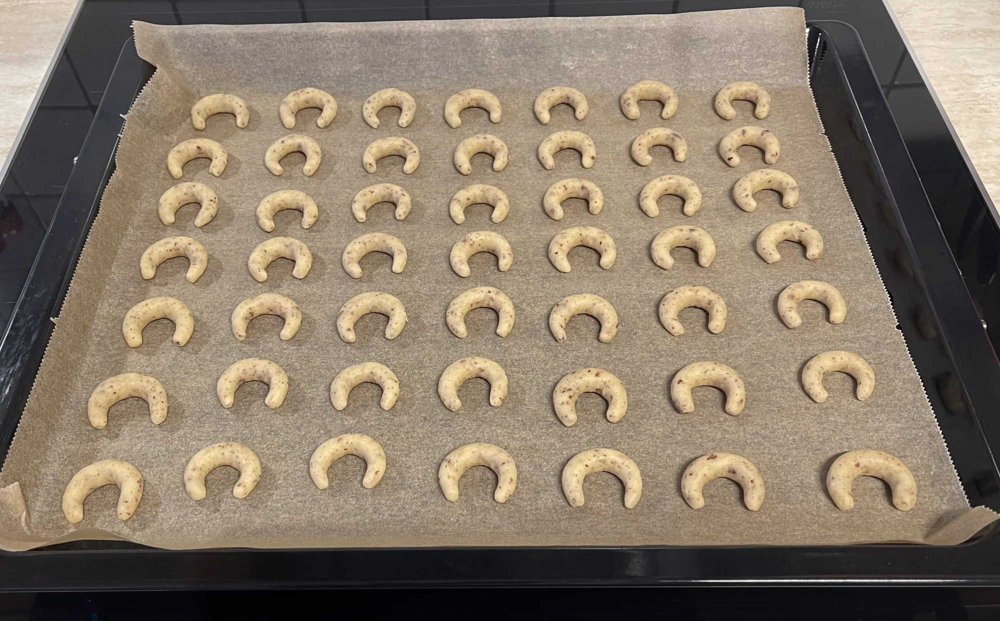 Ungebackene Vanillekipferl mit Nussstückchen in sieben ordentlichen Reihen auf einem mit Backpapier belegten schwarzen Blech