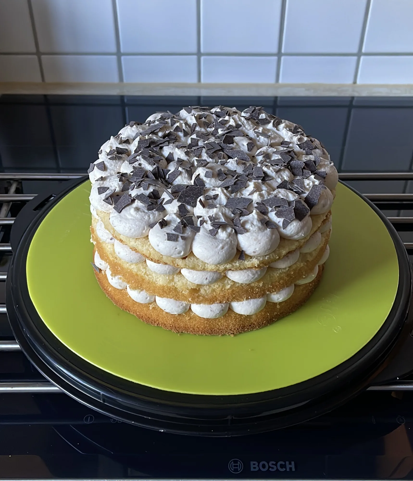 Kleine Torte mit drei Biskuitböden, Sahnetupfen dazwischen und Schokoraspeln obenauf, auf grüner Platte am Herd.