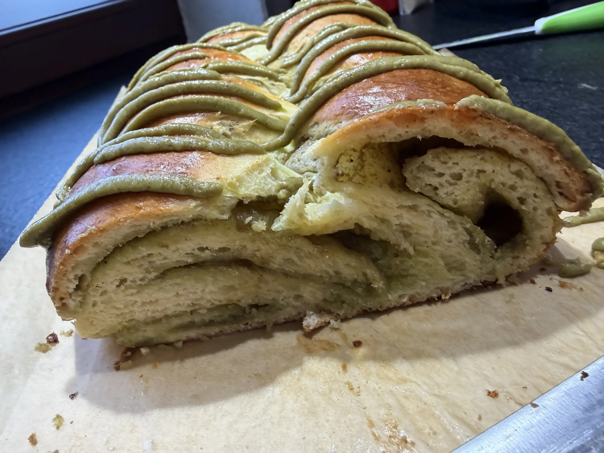 Angeschnittener Hefezopf mit grüner Pistaziencreme-Glasur, im Anschnitt sieht man gefüllte Teigschichten, auf Backpapier.