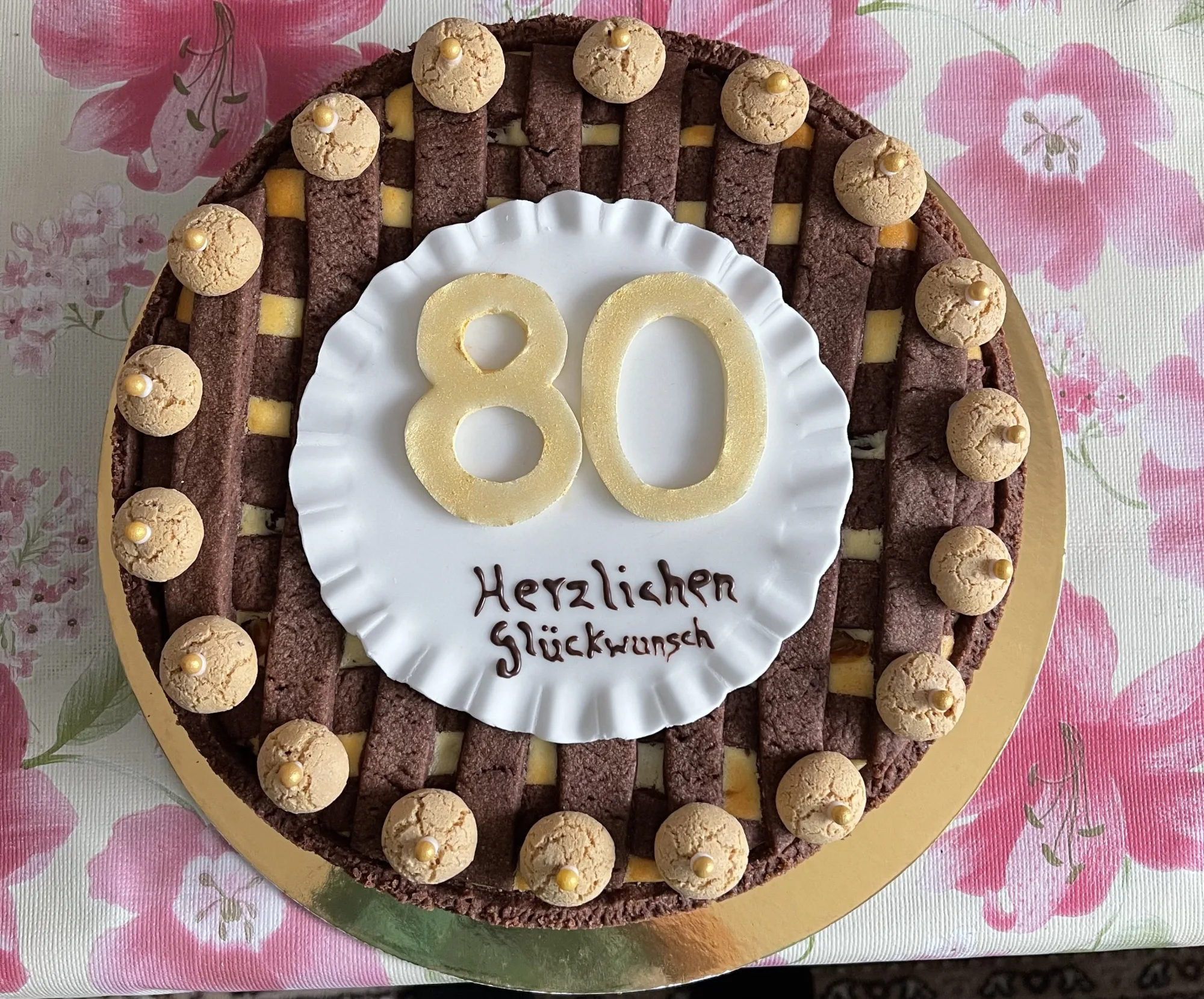 Schokoladen-Gitterkuchen mit Käsefüllung, weißem Zierteller mit goldener „80“, Schriftzug „Herzlichen Glückwunsch“ und Amarettini-Kranz