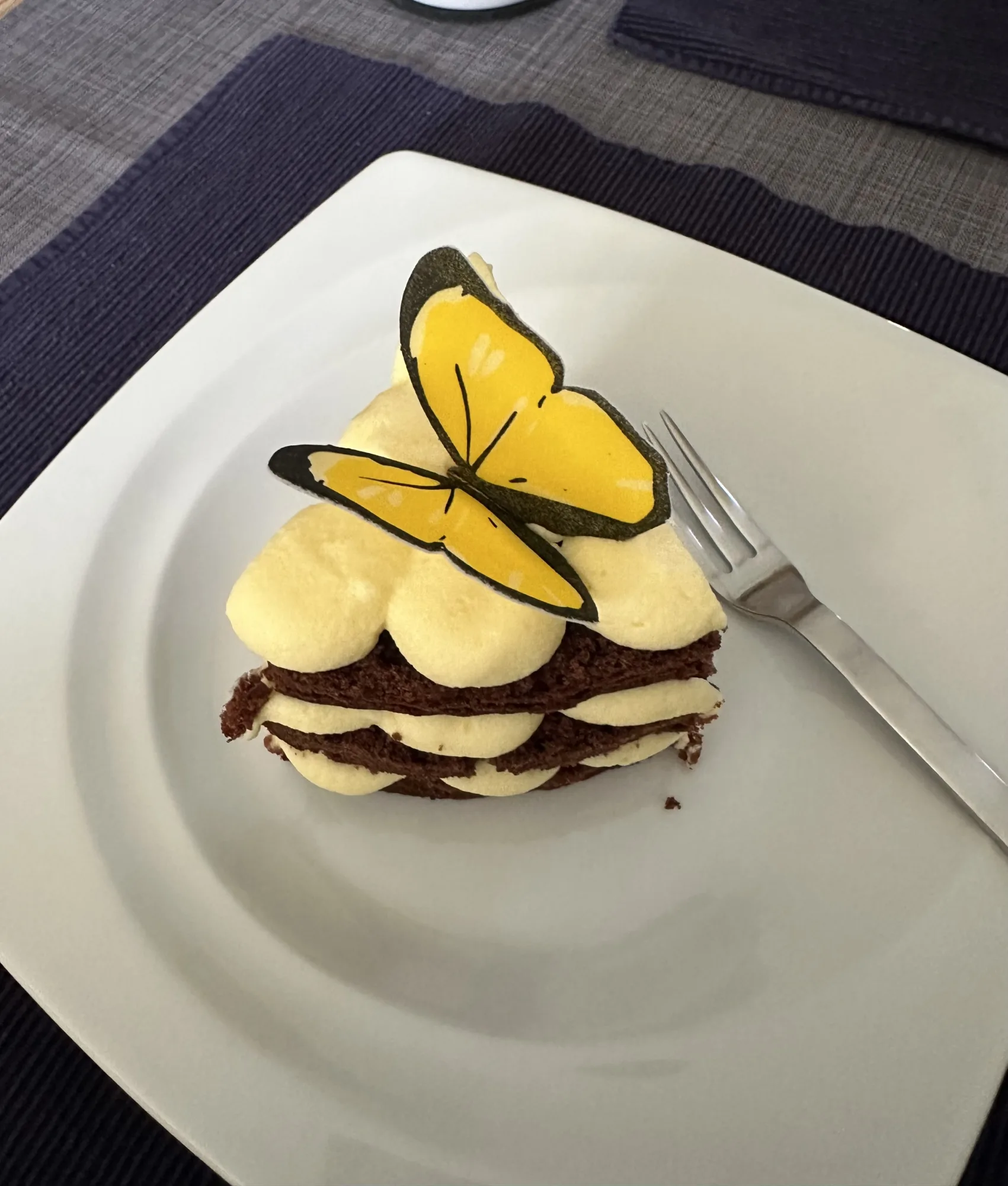 Kleine Torte aus dunklen Biskuitschichten mit gelber Cremetupfen und gelbem Schmetterling aus Oblate, auf weißem Teller mit Gabel