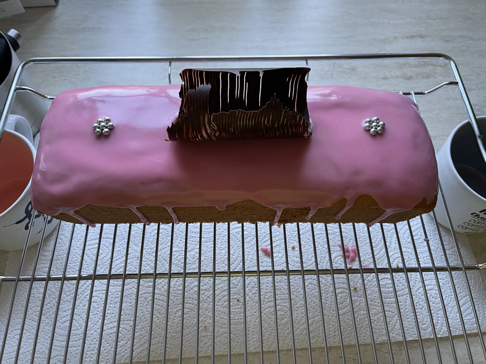 Kastenkuchen mit rosa Zuckerguss, zwei silbernen Perlenblümchen und einem Schokoladenstück obenauf, abkühlend auf einem Gitter