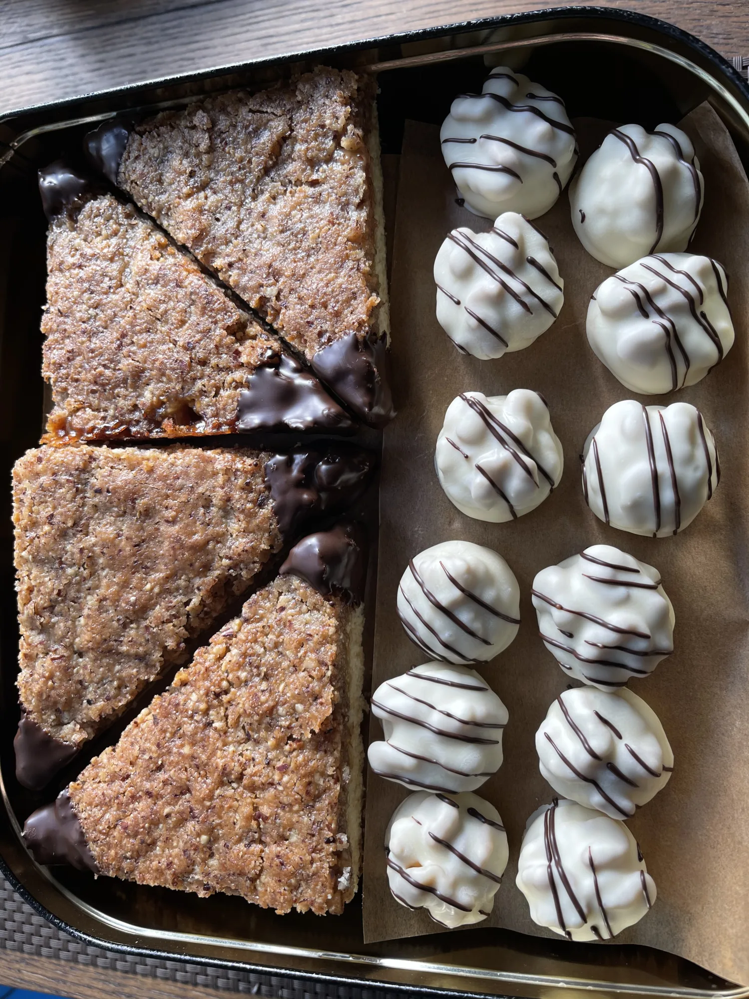 Nussecken mit dunkel getauchten Spitzen neben weißen Schokohäufchen mit dunklen Streifen, in einer Blechdose auf Backpapier