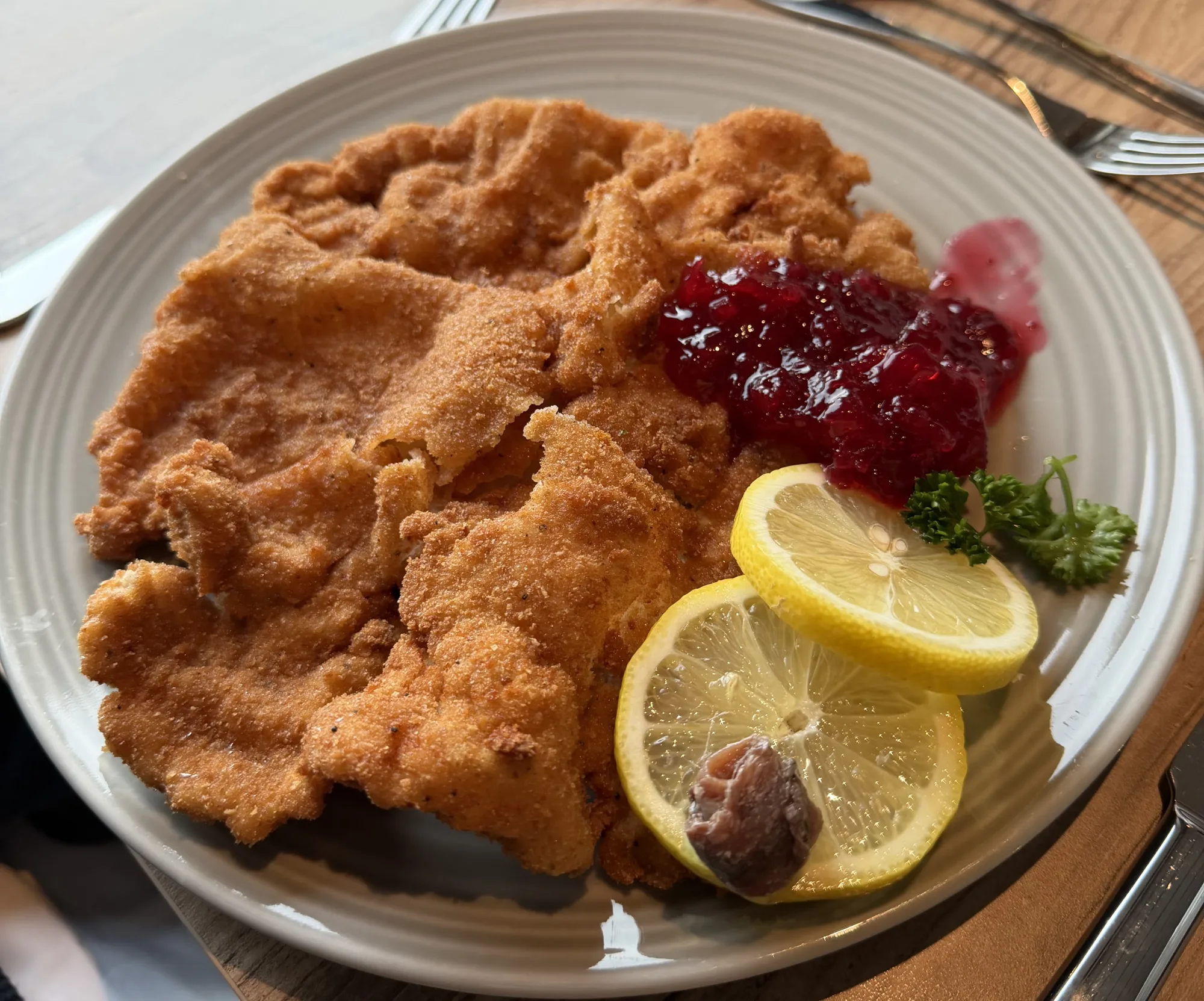 Wiener Schnitzel with wavy golden-brown breading on a gray plate, beside lingonberry jam, two lemon slices with anchovy, and parsley