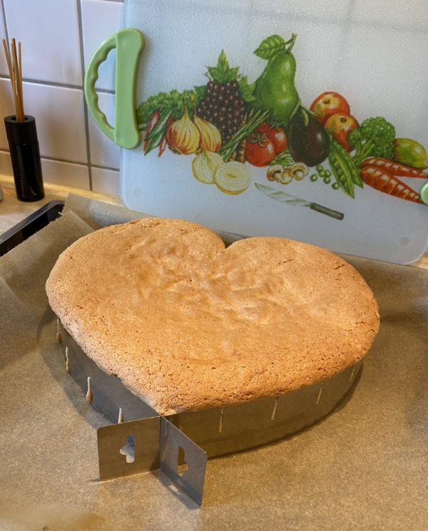 Herzförmiger Biskuitboden mit goldbrauner Oberfläche im verstellbaren Backrahmen auf Backpapier, dahinter ein Gemüse-Schneidbrett.