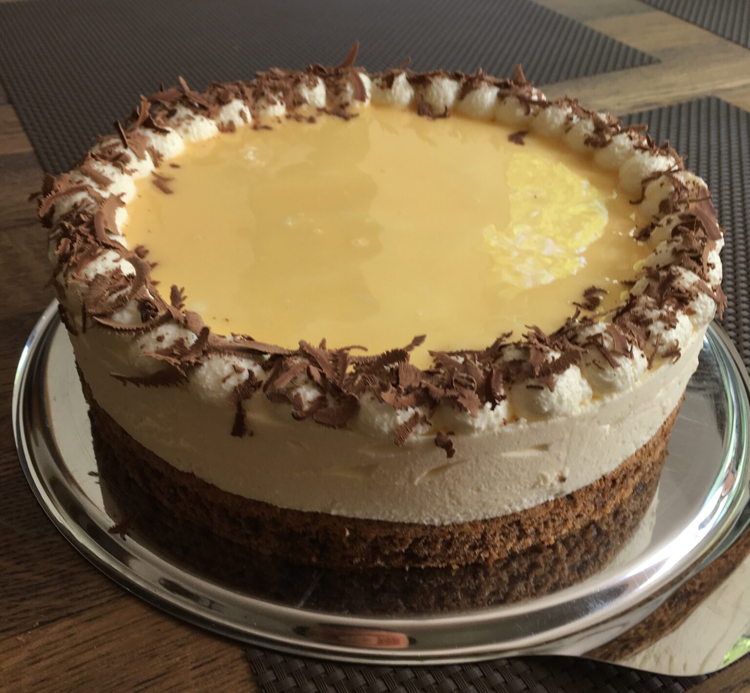 Egg liqueur cream cake with dark base, pale cream layer, yellow glaze, cream dollops and chocolate shavings on a metal platter