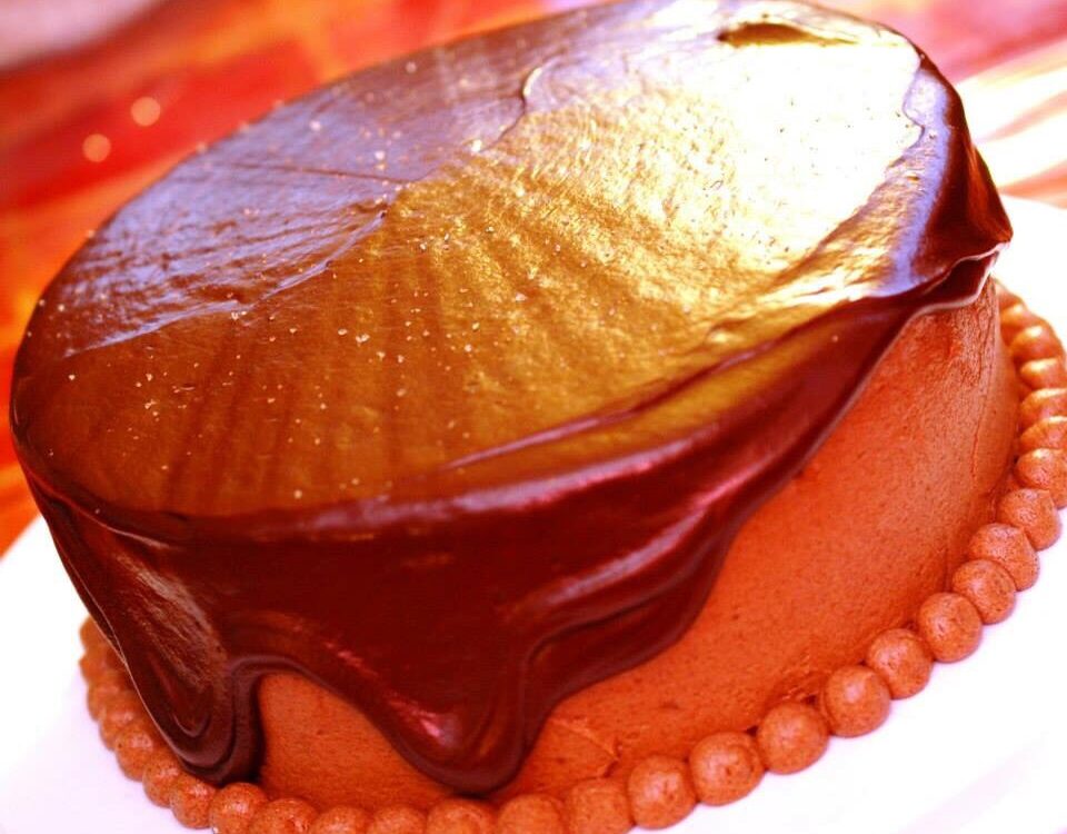 Kleine runde Torte mit glänzender Vollmilch-Ganache oben, die an den Seiten herunterläuft, unten ein Rand aus Cremetupfen auf weißem Teller