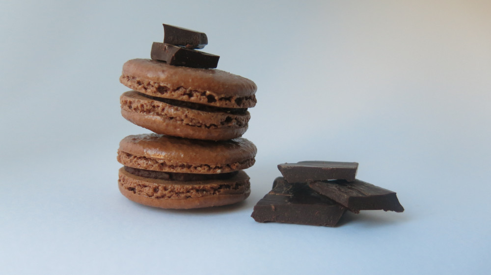 Zwei gestapelte Schoko-Macarons mit dunkler Ganache-Füllung, obenauf Zartbitterschokolade, daneben weitere Schokoladenstücke.