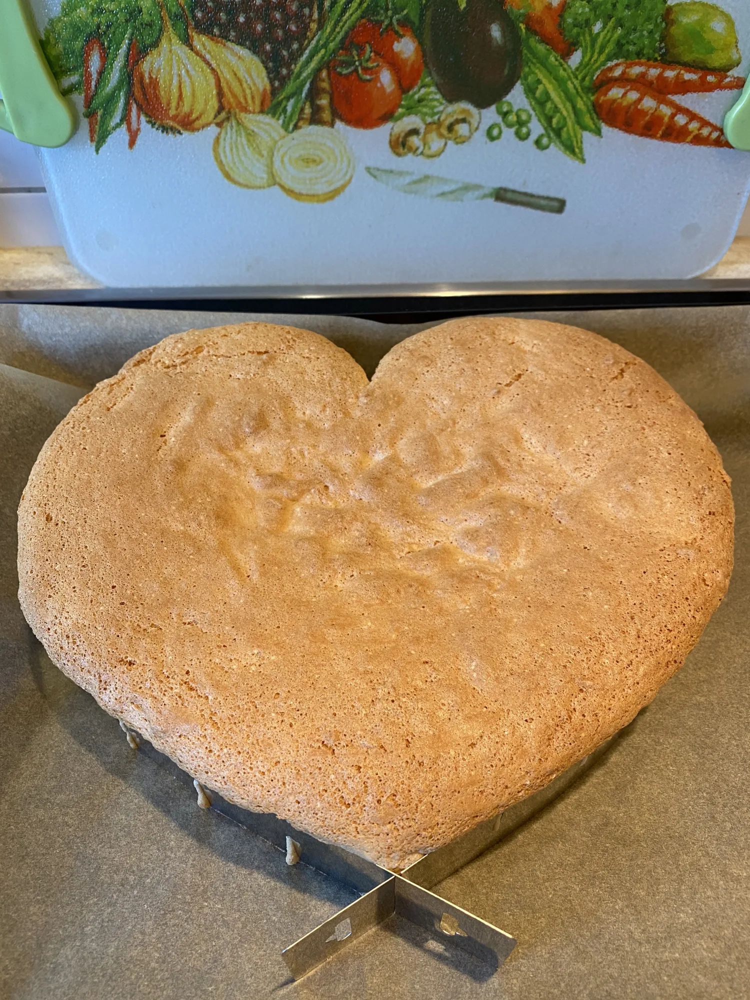 Heart-shaped sponge cake base with a golden-brown top in a metal ring on baking paper, a vegetable-print board behind it