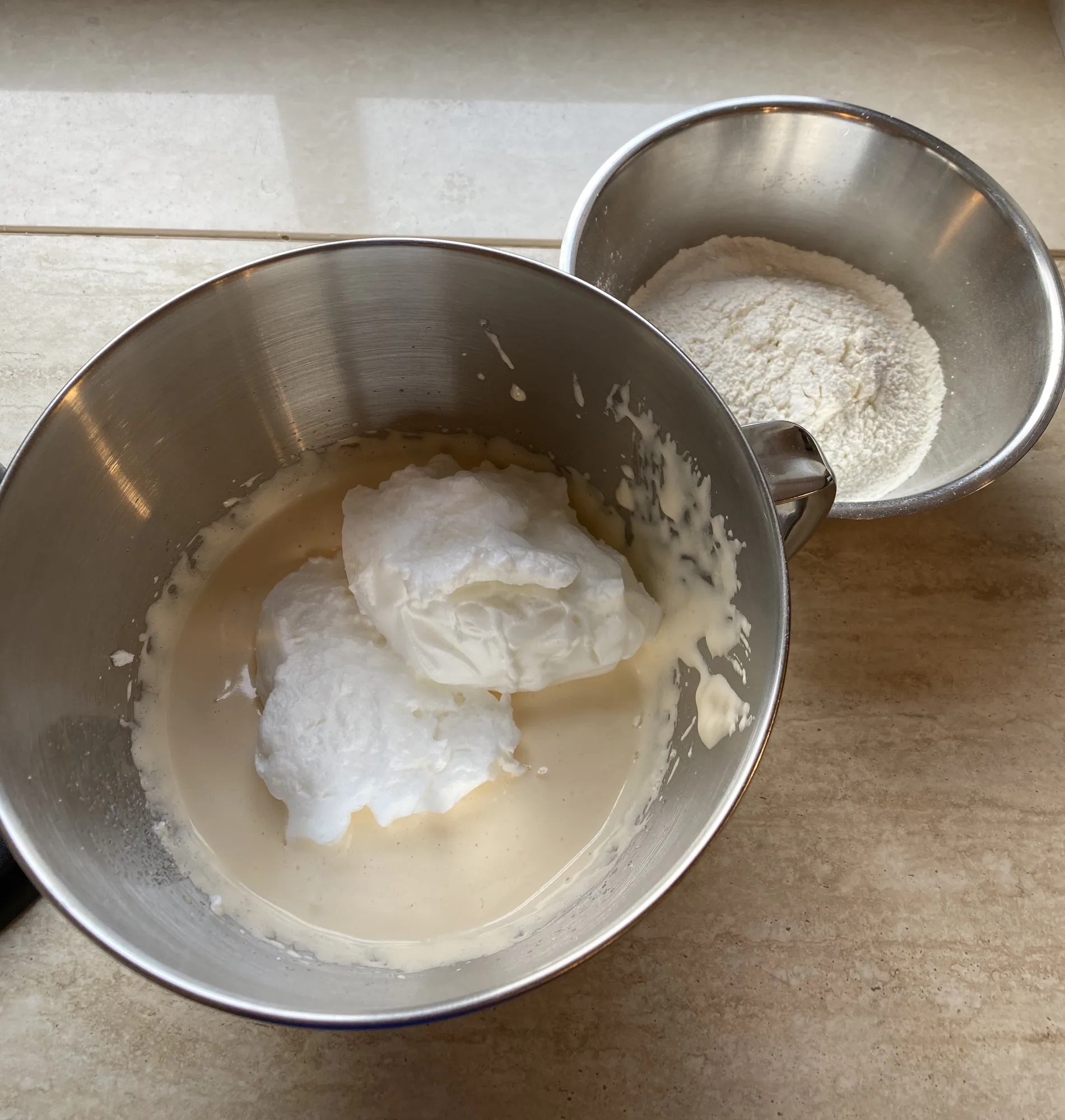 Sponge cake base: beaten egg whites rest on a creamy yolk mixture in a steel mixing bowl, with a bowl of flour beside it.