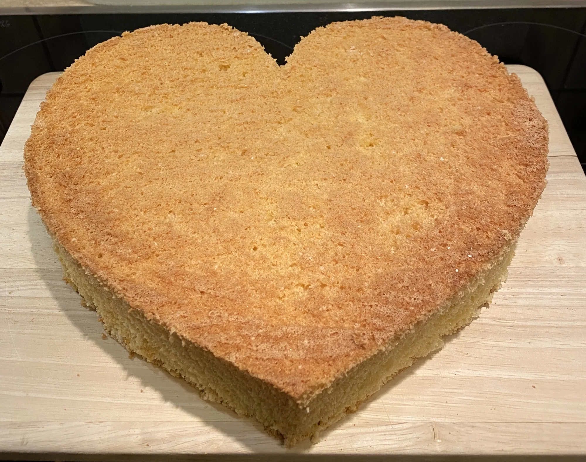 Heart-shaped sponge cake base with a golden-brown, fine-pored top on a light wooden board, unfilled and undecorated.