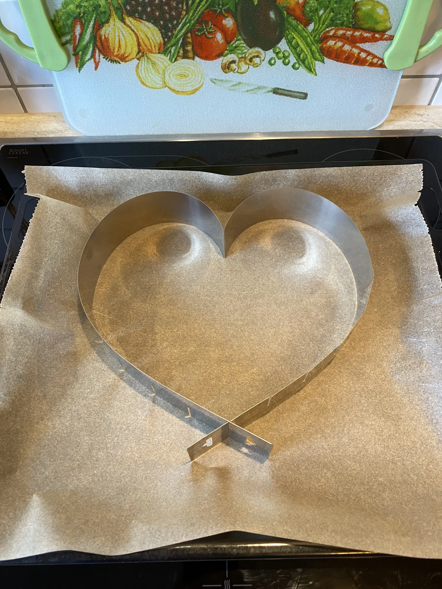 Heart-shaped stainless steel cake ring on parchment paper, ready for the sponge cake base, on a baking tray on the stovetop.