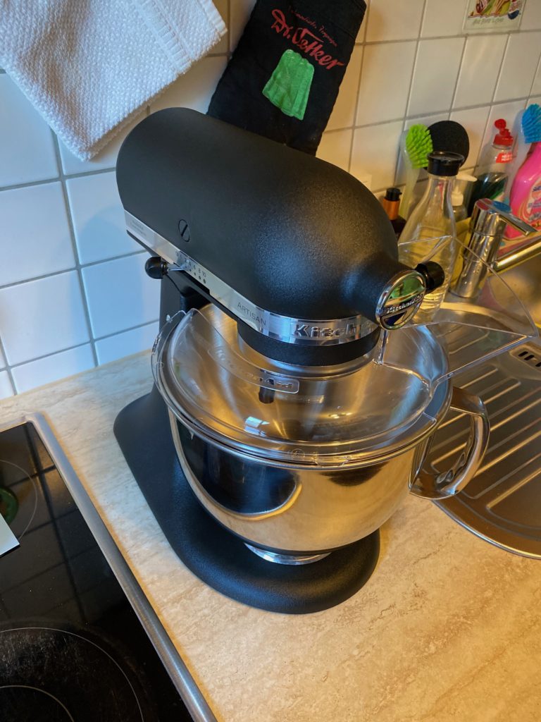 Schwarze KitchenAid Artisan Küchenmaschine mit Edelstahlschüssel und durchsichtigem Spritzschutz auf der Arbeitsplatte neben der Spüle
