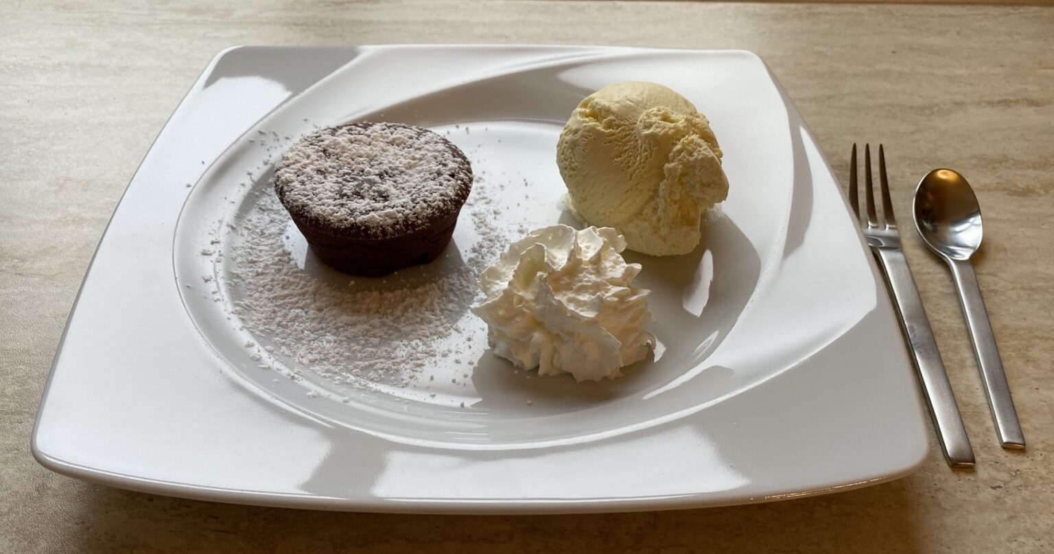 Schokosoufflé mit Puderzucker auf weißem Teller, daneben eine Kugel Vanilleeis und ein Sahnetupfer, rechts Gabel und Löffel