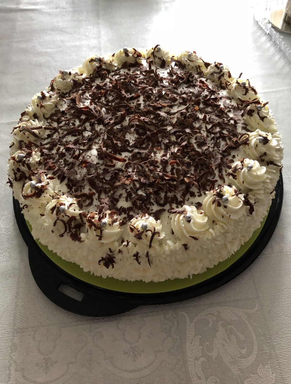 Black Forest cake with cream rosettes, silver sugar pearls and chocolate shavings on a black tray atop a light tablecloth